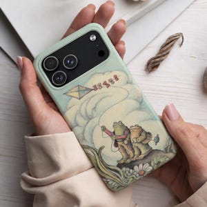 May include: A light green smartphone case featuring a whimsical illustration of two frogs flying a kite in a cloudy sky. The design includes detailed line work, soft pastel colors, and a small daisy flower at the bottom, covering the back of the device.