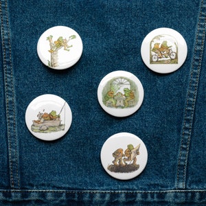 May include: Five round white buttons with images of frogs on them. The frogs are depicted in various activities, including riding a bicycle, fishing, sitting at a table, and climbing a tree.