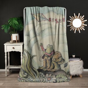 May include: A throw blanket featuring a whimsical illustration of two frogs flying a kite. The frogs are green and brown with red accents. The kite is blue and white with a red ribbon tail. The background is a watercolor wash of blue and green.