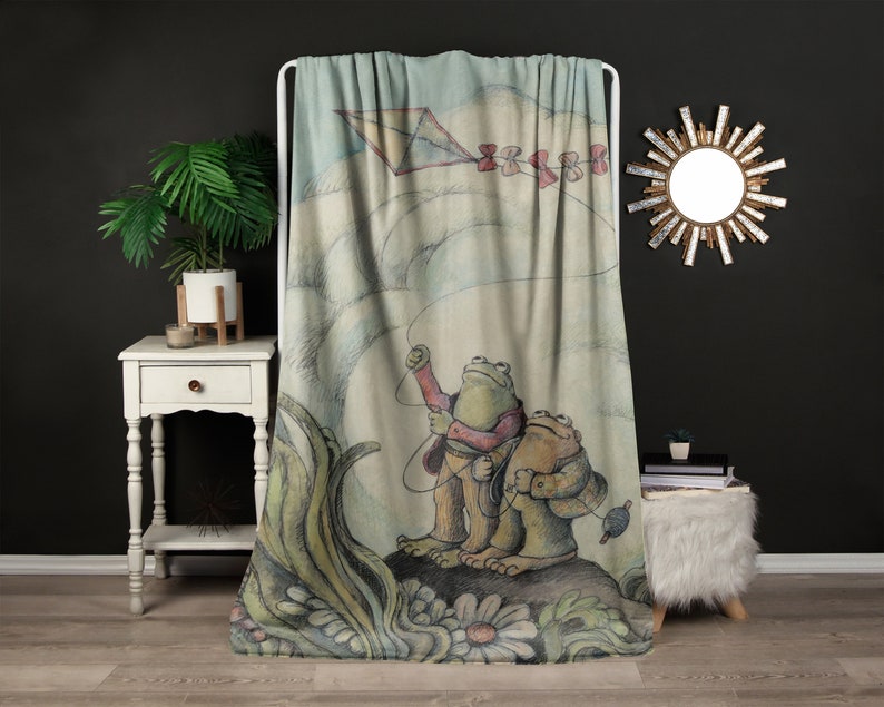 May include: A throw blanket featuring a whimsical illustration of two frogs flying a kite. The frogs are green and brown with red accents. The kite is blue and white with a red ribbon tail. The background is a watercolor wash of blue and green.
