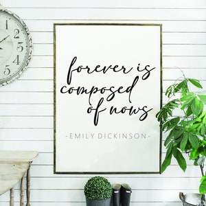 May include: A framed print with a white background and black text that reads "forever is composed of nows -EMILY DICKINSON-". The print is hanging on a white wall with wood paneling. There is a clock, a table, a plant, and a pair of boots in the image.