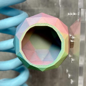 May include: A multi-colored, geometric-shaped object with a hollow interior. The object has a faceted design with pastel colors including pink, green, and blue. The object is near a light blue spiral and a clear surface with metal fasteners.
