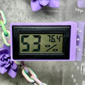 Może przedstawiać: Cyfrowy higrometr z czarnym wyświetlaczem i fioletową ramką. Wyświetlacz pokazuje poziom wilgotności 53% i temperaturę 24,7°C. Urządzenie jest połączone z łańcuchem.