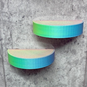 Puede incluir: Dos estantes semicirculares con un degradado de tonos verdes, azules y rosas. Los estantes están montados en una pared gris texturizada. Los estantes parecen estar hechos de un material impreso en 3D.