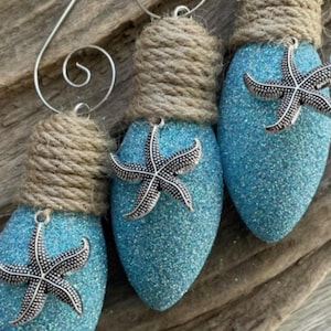 May include: Three blue lightbulb ornaments decorated with silver starfish and wrapped with twine. The ornaments are hanging from a rustic wooden surface.