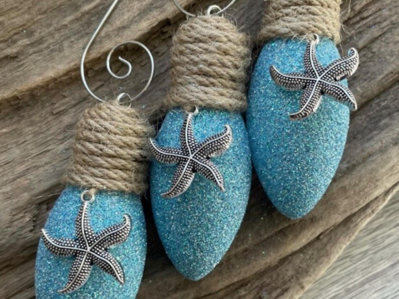 May include: Three blue lightbulb ornaments decorated with silver starfish and wrapped with twine. The ornaments are hanging from a rustic wooden surface.