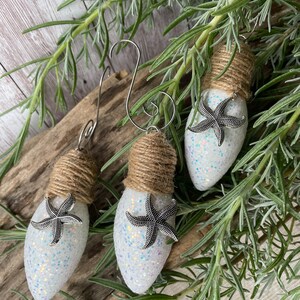 May include: Three white lightbulb ornaments with silver glitter and a silver starfish attached to each. The ornaments are wrapped with twine and have a silver wire hanger.
