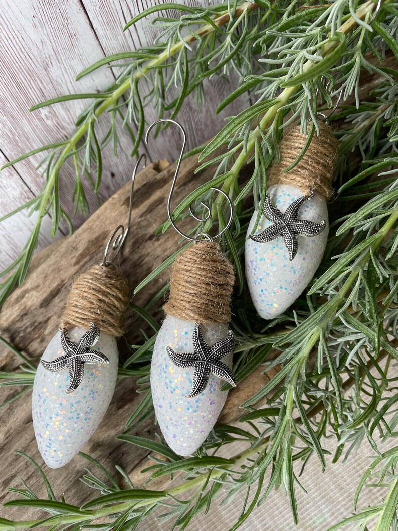 May include: Three white lightbulb ornaments with silver glitter and a silver starfish attached to each. The ornaments are wrapped with twine and have a silver wire hanger.