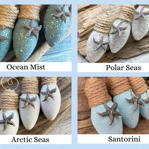 May include: Four sets of lightbulb ornaments decorated with twine and glitter. Each set has a different color scheme: Ocean Mist (blue), Polar Seas (white), Arctic Seas (white), and Santorini (blue). Each ornament features a silver starfish.