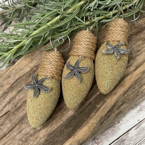 May include: Three light bulb ornaments decorated with burlap and glitter, each with a silver starfish attached. The ornaments are a light green color and have a rustic, beachy feel.