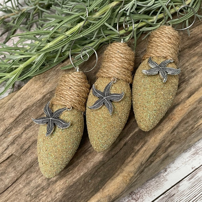 May include: Three light bulb ornaments decorated with burlap and glitter, each with a silver starfish attached. The ornaments are a light green color and have a rustic, beachy feel.