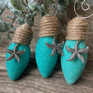May include: Three teal blue lightbulb ornaments with glitter and silver starfish accents. Each ornament is topped with a brown twine bow and a silver wire hanger.