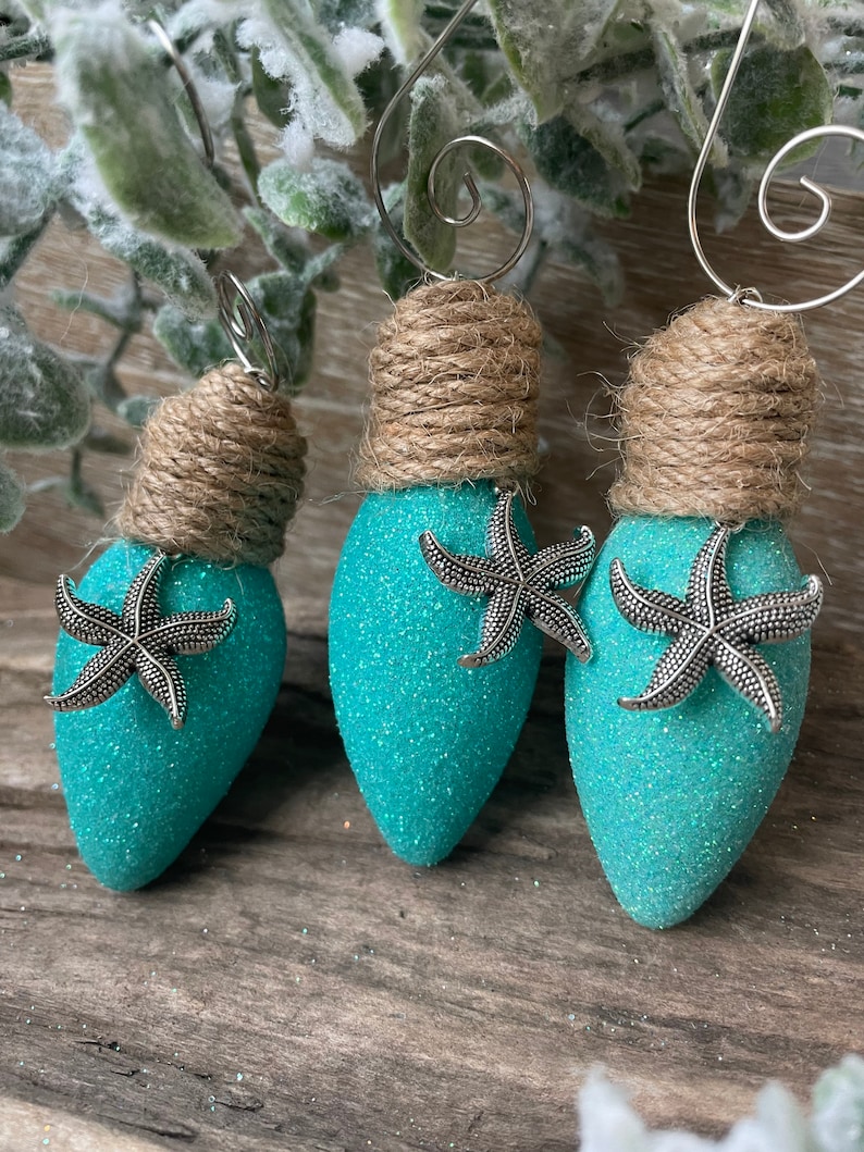 May include: Three teal blue lightbulb ornaments with glitter and silver starfish accents. Each ornament is topped with a brown twine bow and a silver wire hanger.