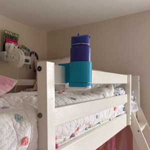 May include: A blue and teal water bottle holder attached to a white bunk bed rail. The water bottle is a dark blue color with a silver band. The bed has a white comforter with colorful circular accents.