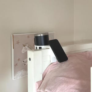 Op de afbeelding: Een zwarte geïsoleerde beker met een zilveren rand staat op een wit bedframe, naast een zwarte smartphone. Een ingelijst eenhoornkunstwerk hangt aan de muur, met een roze deken op het bed.