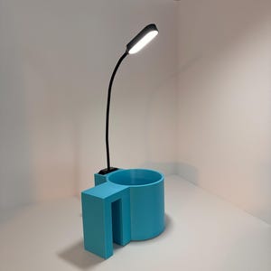May include: A modern desk lamp with a flexible black neck and a rectangular light head. The lamp is attached to a light blue, geometric-shaped base with a cylindrical cup holder. The lamp is turned on.