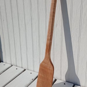 May include: A long, light brown wooden spatula with a smooth, flat, rectangular head and a rounded handle. The grain of the wood is visible throughout the surface of the utensil, which is positioned vertically against a white paneled surface.