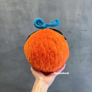 May include: An orange plush fruit with flame-like patterns and a blue, key-shaped top. The plush toy has a black band around the base and the text "Chalkshock" is visible.