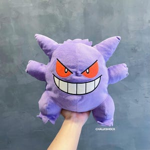 Puede incluir: Juguete de peluche morado de un Pokémon fantasma con una sonrisa amenazante y ojos rojos. El juguete está siendo sostenido por una mano.