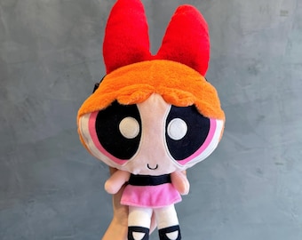 May include: A Blossom plush toy from The Powerpuff Girls. The doll has a large head with orange hair, a red bow, and big eyes. She is dressed in a pink dress, black shoes, and has a small smile. The doll is held against a grey background.