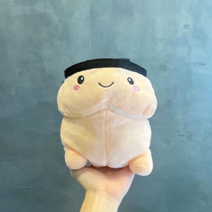 Puede incluir: Una bolsa de cordón de felpa rosa y negra con una cara sonriente y mejillas rosadas. La bolsa está hecha de un material suave y peludo y tiene un cierre de cordón negro.