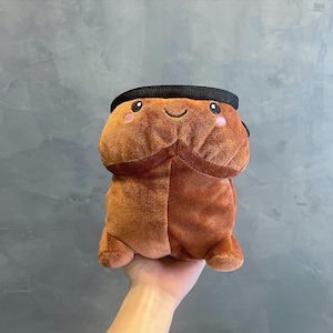 Puede incluir: Un juguete de peluche marrón con una cara sonriente, mejillas rosadas y una banda negra alrededor de la parte superior. El juguete tiene una forma única y está hecho de material suave.