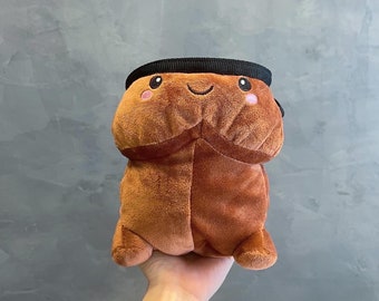 May include: A brown plush toy with a smiling face, pink cheeks, and a black band around the top. The toy has a unique shape and is made of soft material.