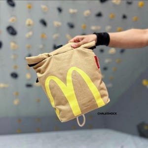 Könnte beinhalten: Ein beigefarbener Chalkbag mit gelbem McDonald's-Logo. Der Beutel hat einen Kordelzugverschluss und ein kleines rotes Etikett. Das Wort "CHALKSHOCK" ist unten rechts im Bild aufgedruckt.