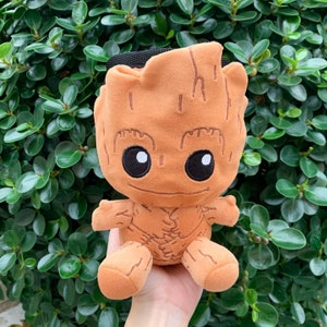 Puede incluir: Un peluche del personaje Groot de la franquicia Guardianes de la Galaxia. El juguete es naranja con ojos negros y una pequeña sonrisa. Tiene una correa negra en la parte superior y se sostiene en una mano sobre un fondo de hojas verdes.