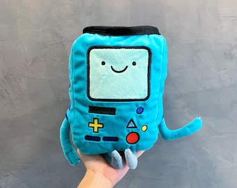 May include: A bright blue plush toy shaped like a rectangular video game console with a light blue screen, a smiling face, and embroidered buttons including a yellow plus sign, a blue triangle, a red circle, and a small yellow circle.