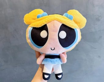 May include: A plush toy of the character Bubbles from The Powerpuff Girls. The doll features a yellow head with two fluffy pigtails, blue bows, and large blue eyes. She is dressed in a light blue dress and white trousers with black shoes.