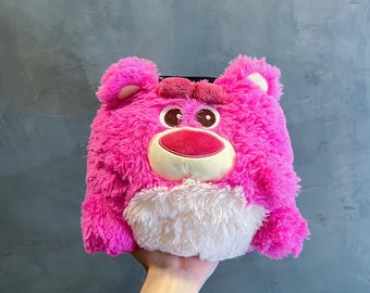 May include: Pink plush toy shaped like a bear with a white face and brown eyes. The bear has a pink nose and a white mouth.