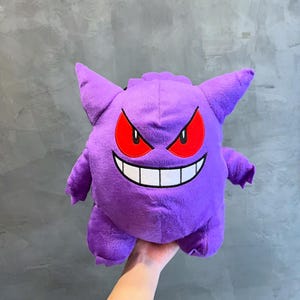 Puede incluir: Juguete de peluche morado de un personaje de Pokemon con una gran sonrisa de dientes y ojos rojos. El personaje es conocido como Gengar.