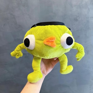 Może przedstawiać: Pluszowa zabawka w kolorze limonki, przypominająca postać z kreskówki, z dużymi białymi i czarnymi oczami, pomarańczową płetwą i małymi kończynami. Ma czarny pasek na górze i jest trzymana w dłoni.