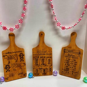May include: Three wooden cutting boards with handles, each engraved with kitchen-related text and graphics. One board shows cooking temperatures for meats, another a conversion chart, and the third a cheat sheet. Pink and white candy-shaped beads hang above.