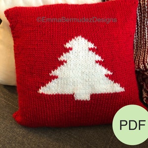 May include: Red knitted pillow with a white knitted Christmas tree design. The pillow has the text "©EmmaBermudezDesigns" on it. The pillow is on a brown couch. A green circle with the text "PDF" is in the lower right corner of the image.