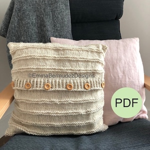 May include: A cream-colored, knitted pillow with horizontal ribbed detailing and wooden buttons. The pillow is part of a home decor set, with a pink pillow and a dark gray blanket in the background. The text "EmmaBermudezDesigns" is visible.