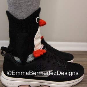 May include: Black and white knitted penguin sock with red accents. The sock is worn on a person's foot with a black and white sneaker. The text "EmmaBermudezDesigns" is visible on the sneaker.