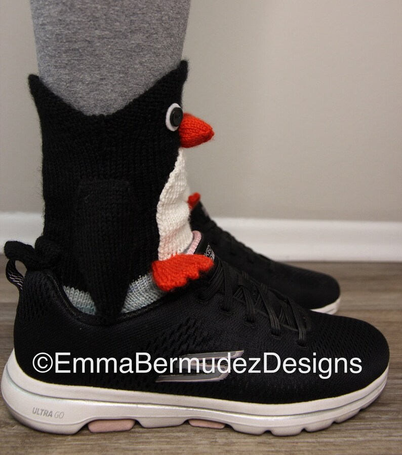 May include: Black and white knitted penguin sock with red accents. The sock is worn on a person's foot with a black and white sneaker. The text "EmmaBermudezDesigns" is visible on the sneaker.