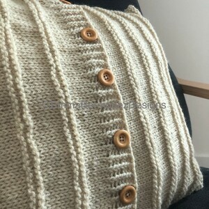 May include: Cream-colored knitted pillow with a ribbed design and wooden buttons. The pillow has a cardigan-like appearance, with vertical ribbing and a button closure. The pillow is on a dark gray chair.