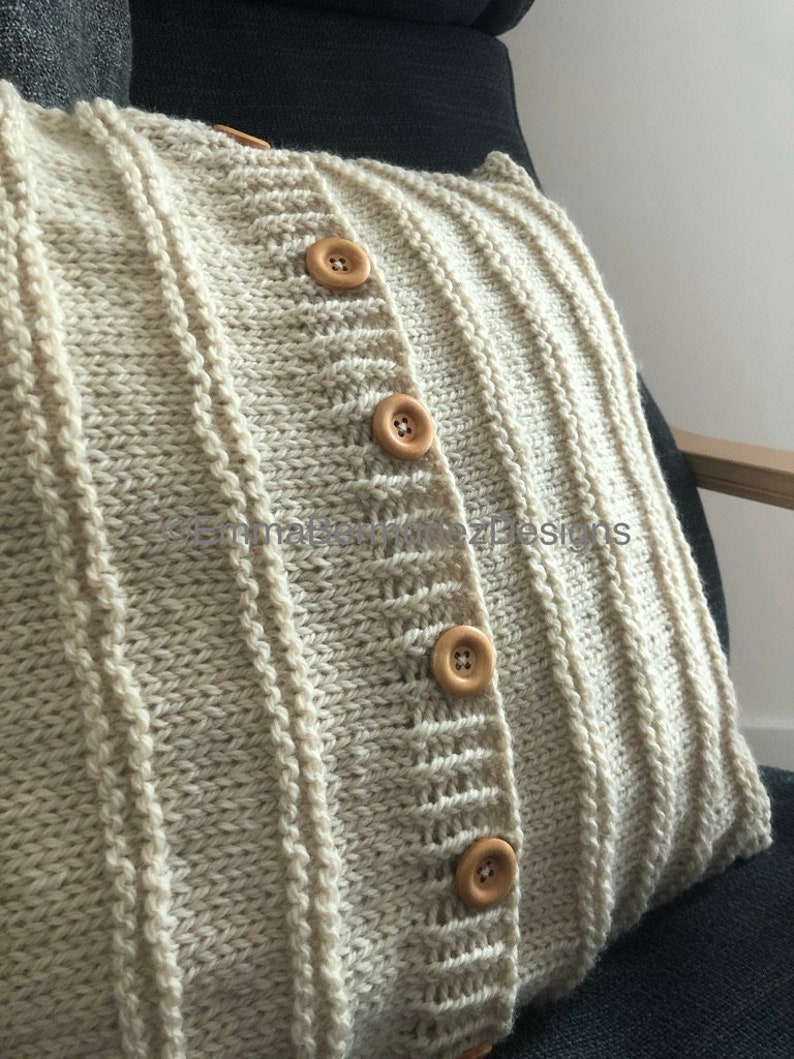 May include: Cream-colored knitted pillow with a ribbed design and wooden buttons. The pillow has a cardigan-like appearance, with vertical ribbing and a button closure. The pillow is on a dark gray chair.