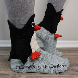 May include: A pair of blue and white knit socks with penguin appliques. The penguins are black with white bellies and orange beaks and feet. The socks are made with a textured knit and have a light blue and white color pattern.