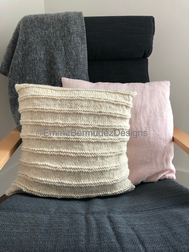 May include: A cream-colored, horizontally ribbed knitted pillow rests on a dark gray chair. A light pink pillow and a gray throw blanket are also on the chair. The chair has a dark gray seat and back.
