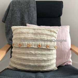May include: A cream-colored, knitted pillow with horizontal ribbed detailing and wooden buttons, resting on a chair with a gray cushion. A gray blanket and a pink pillow are also on the chair. The chair has wooden arms.