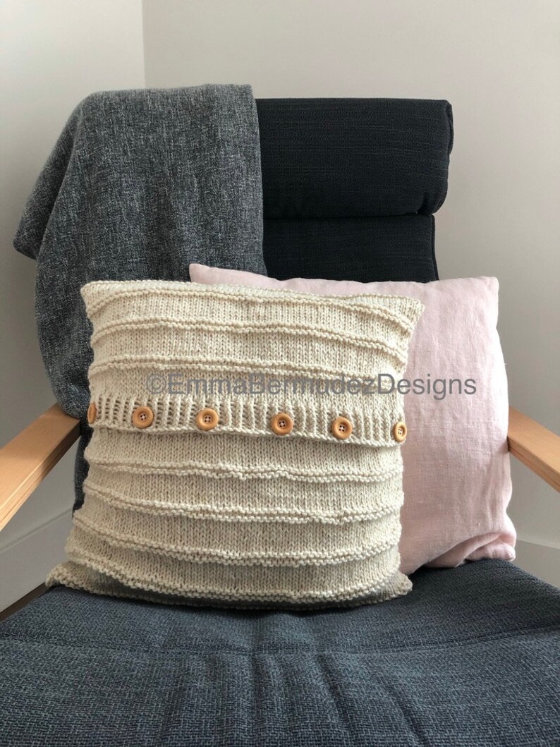 May include: A cream-colored, knitted pillow with horizontal ribbed detailing and wooden buttons, resting on a chair with a gray cushion. A gray blanket and a pink pillow are also on the chair. The chair has wooden arms.