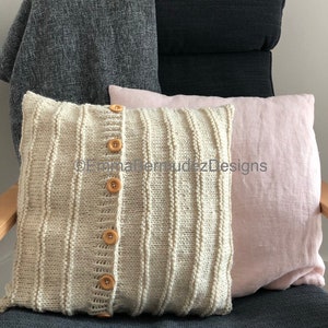 May include: A decorative pillow with a knitted front, featuring vertical ribbing and wooden buttons. The pillow is cream-colored and has a light pink linen pillow next to it. A gray blanket is draped over the back of a dark gray chair.