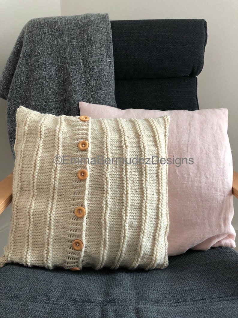 May include: A decorative pillow with a knitted front, featuring vertical ribbing and wooden buttons. The pillow is cream-colored and has a light pink linen pillow next to it. A gray blanket is draped over the back of a dark gray chair.