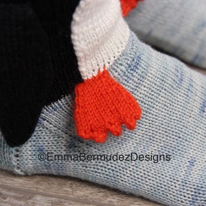 May include: A pair of blue knitted socks with a white and orange penguin design. The penguin has a black beak and orange feet. The socks are made of a soft, comfortable material.
