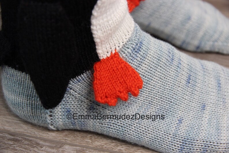 May include: A pair of blue knitted socks with a white and orange penguin design. The penguin has a black beak and orange feet. The socks are made of a soft, comfortable material.
