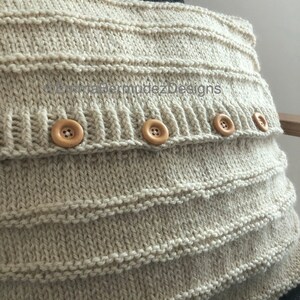 May include: A beige knitted pillow cover with horizontal ribbed detailing and four wooden buttons. The pillow cover is made of a soft, textured yarn and has a simple, classic design. The text "BermudezDesigns" is visible on the pillow cover.